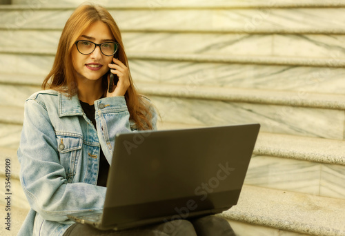 All in business. Young stylish woman in a denim jacket and glasses uses a laptop and talking on the phone while sitting on the stairs in the city. Remote work.