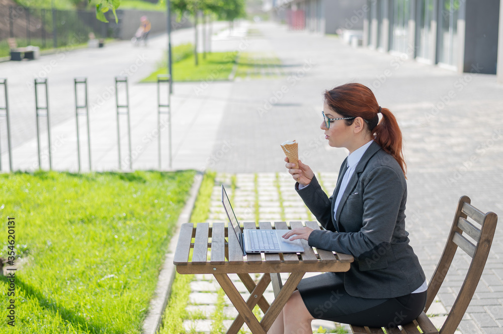 Fototapeta premium Happy woman typing on the computer and eating ice cream cone while sitting at a wooden table outdoors. Female employee remotely working on a laptop. Lunch break in a street cafe.