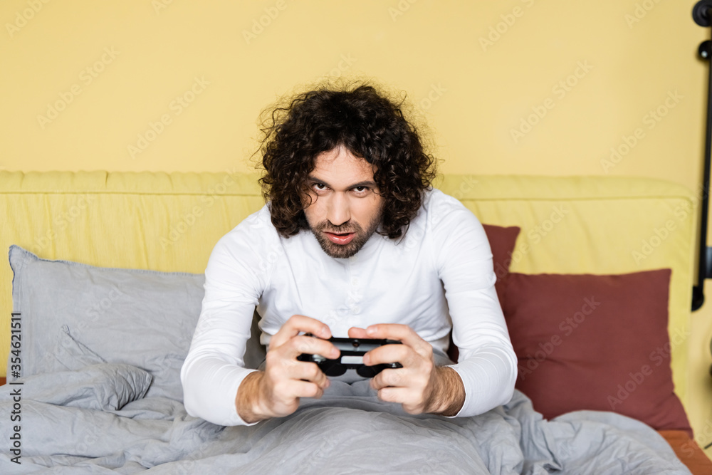 KYIV, UKRAINE - APRIL 25, 2020: concentrated, curly man playing video game with joystick in bedroom KYIV, UKRAINE - APRIL 25, 2020: concentrated, curly man playing video game with joystick in bedroom