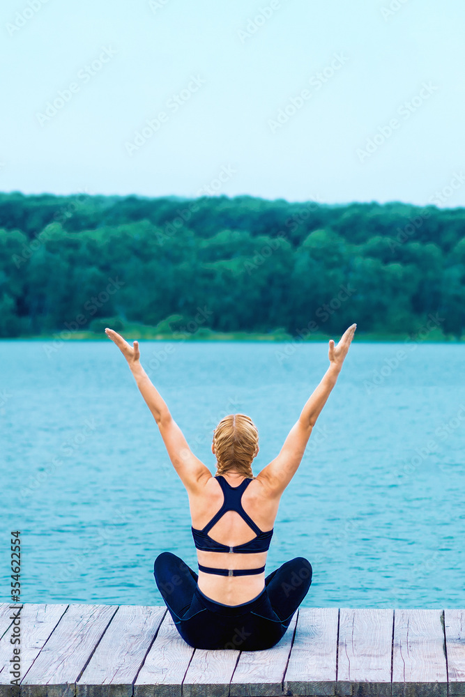 Back view of young woman is doing yoga exercise on grass of the shore of the lake.