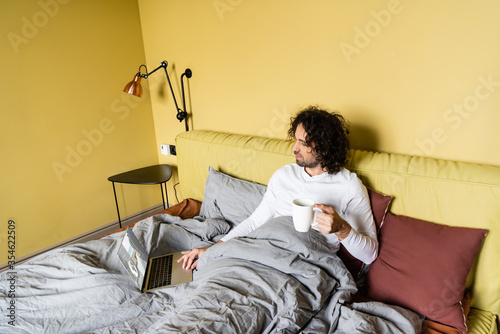 high angle view of young freelancer using laptop with tickets online website while holding cup of coffee in bed