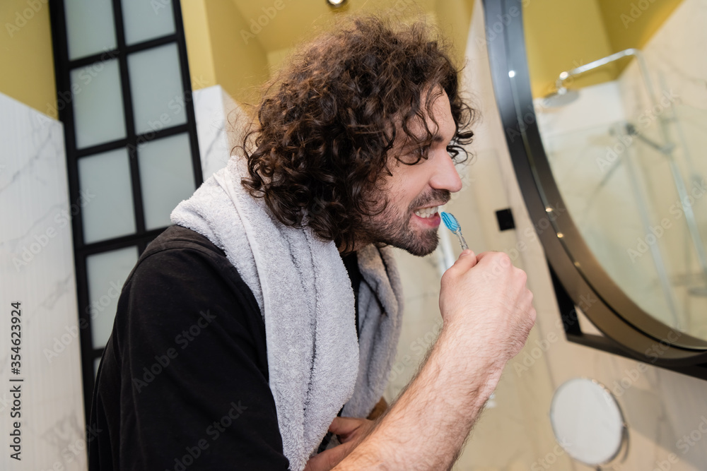 Side view of curly man brushing teeth near mirror in bathroom