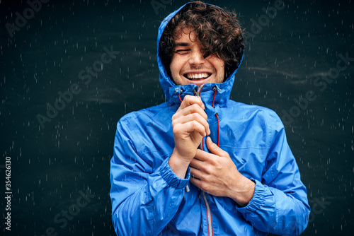 Portrait of the man smiling broadly, wearing blue raincoat during rain outside. Handsome male in blue raincoat enjoying the rain on black wall. Young man has joyful expression in rainy weather. photo