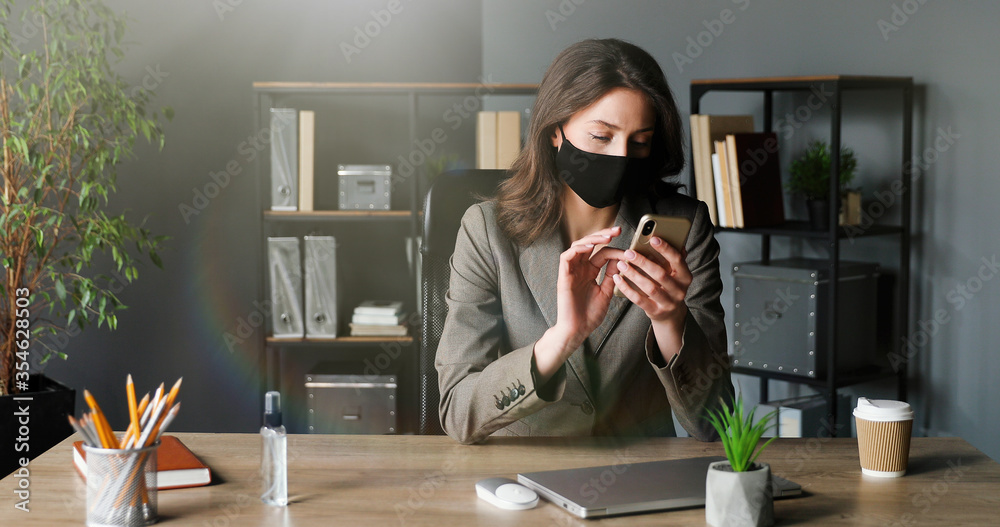 Young businesswoman sitting and talking on cellphone. Beautiful female office worker in mask speaking on mobile phone in cabinet. Coronavirus pandemic time.