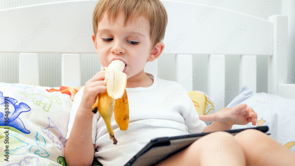 Portrait of little toddler boy watching video on digital tablet and eating banana for breakfast Portrait of little toddler boy watching video on digital tablet and eating banana for breakfast