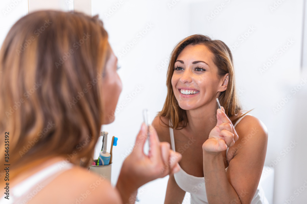 Self care and make up concept. Waist up portrait of young careful woman in white shirt plucking her eyebrows by tweezers before mirror in bathroom Self care and make up concept. Waist up portrait of young careful woman in white shirt plucking her eyebrows by tweezers before mirror in bathroom