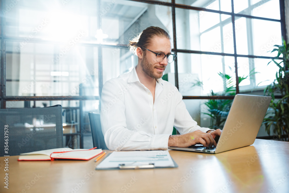 Photo of handsome young man working with laptop while sitting at table Photo of handsome young man working with laptop while sitting at table