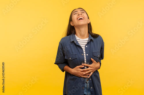 What a funny joke! Portrait of amused carefree girl in denim shirt cracking up with hilarious laughter, holding her stomach and laughing out loud. indoor studio shot isolated on yellow background