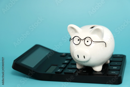 White ceramic piggy bank wearing reading glasses standing on calculator on blue teal background. Concept for money savings plan for retirement, aged society, or financial accounting