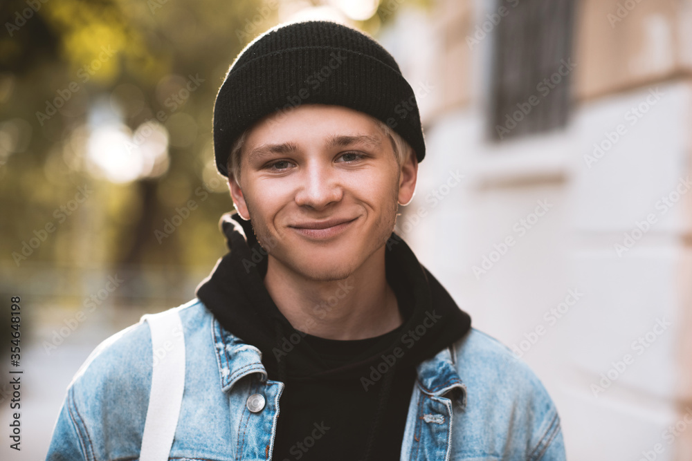 Smiling teen boy 16-18 year old wearing black knitted hat, hoodie and denim jacket outdoors at sun set on background. Looking at camera. Teenagerhood.