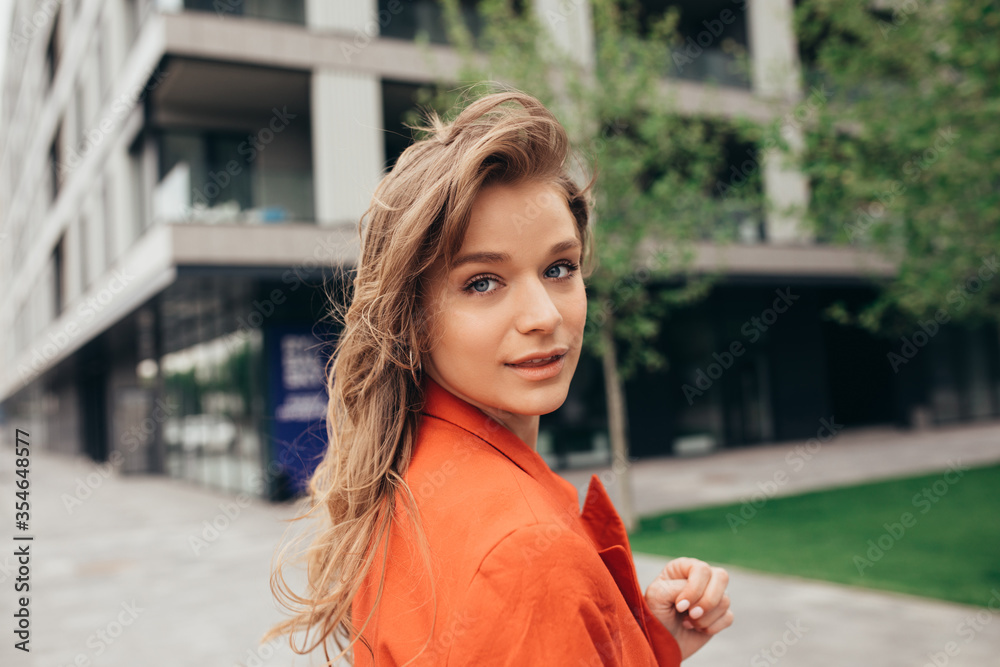 Picture of beautiful stylish young woman look back on camera. Posing alone outside at building. Stylish trendy girl in orange jacket with beautiful face. Street style.
