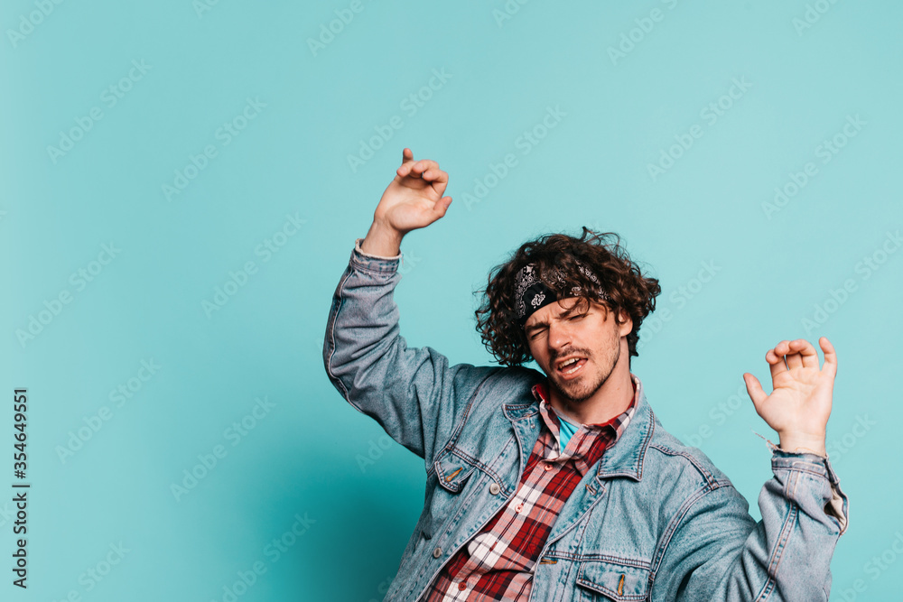 Young positive cheerful hipster isolated over blue background. Guy dancing alone in studio with raised hands up. Having fun during modern dance. Young positive cheerful hipster isolated over blue background. Guy dancing alone in studio with raised hands up. Having fun during modern dance.