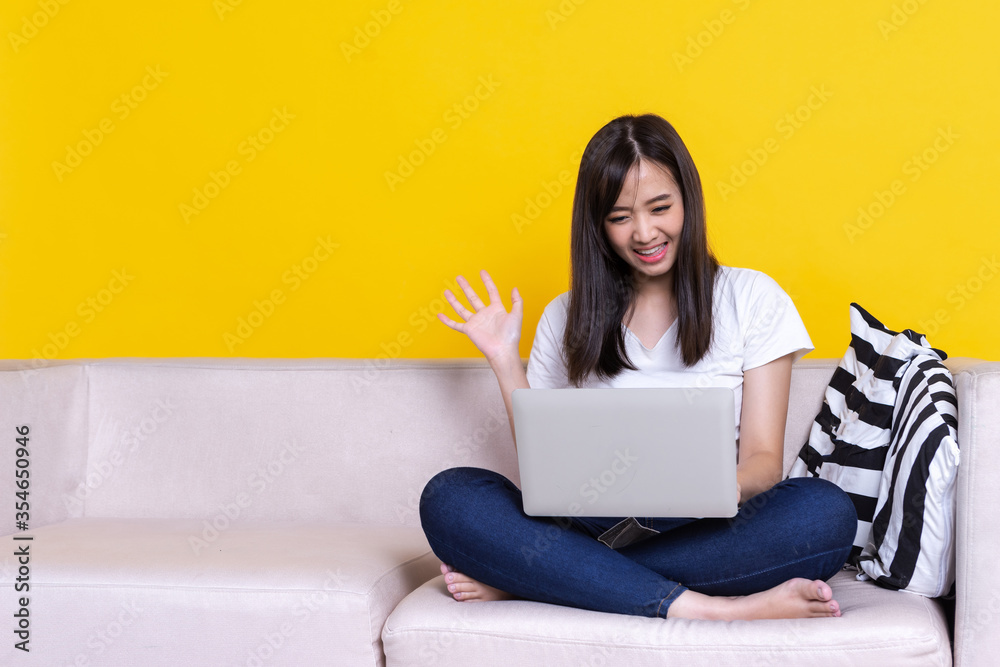 Asian woman smiling and working from home use laptop computer while sitting on sofa over isolate yellow background Asian woman smiling and working from home use laptop computer while sitting on sofa over isolate yellow background