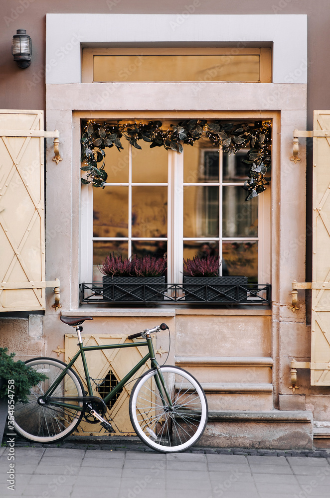 Old white wooden window with rectangular frames for glass and christmas garland with branches with a flower pot with flowers and old-fashioned bike near the beige facade.