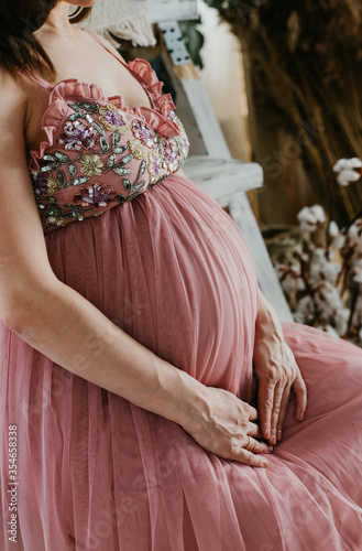 Pregnant woman in dress holds hands on belly. Pregnancy, maternity, preparation and expectation concept. Close-up, copy space, intereior. Beautiful tender mood photo of pregnancy. photo
