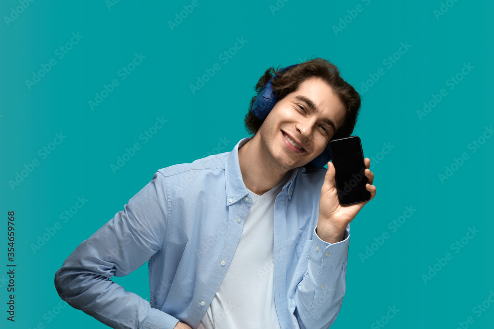 Look at this. Portrait of a young beautiful man wearing white t-shirt and blue shirt in blue headphones holding mobile phone in hand and showing his screen Look at this. Portrait of a young beautiful man wearing white t-shirt and blue shirt in blue headphones holding mobile phone in hand and showing his screen