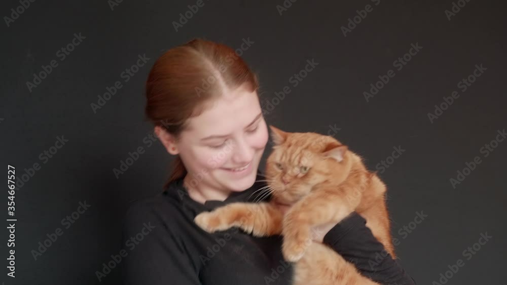 Young caucasian girls holds her red british cat and cuddles it. Black background. Contrast of black and orange. Young caucasian girls holds her red british cat and cuddles it. Black background. Contrast of black and orange.