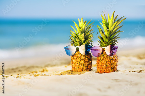 Couple of attractive pineapples in stylish colored sunglasses on the sand against turquoise sea. Tropical summer vacation concept. Sunny day on the beach of tropical island. Honeymoon. Family holiday
