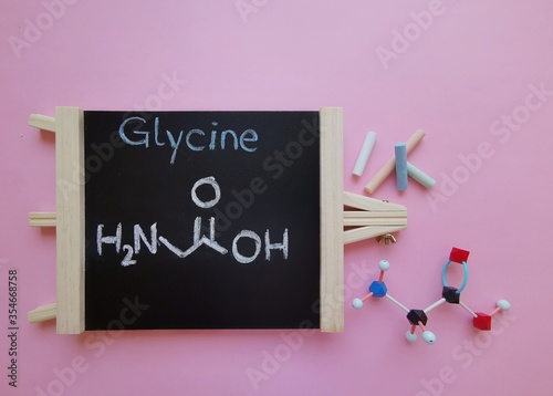 Molecular structure model of glycine molecule with structural chemical formula on a black chalkboard. Glycine (Gly or G) is a proteinogenic amino acid. Black=C, red=O, blue=N, white=H.