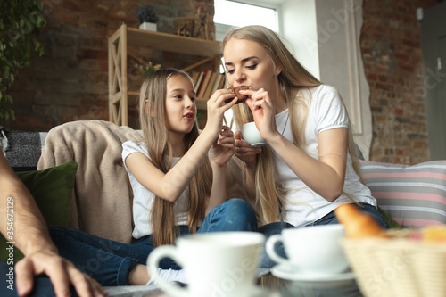 Mother, father and daughter at home having fun, comfort and cozy concept. Looks happy, cheerful and joyful. Beautiful caucasian family. Spending time together, eating cakes, drinking tea on sofa.