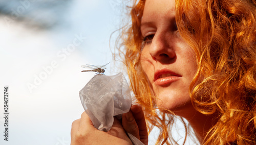 Beautiful red-haired girl is fascinated by the dragonfly, which has landed on the cloth in your hands, copy space