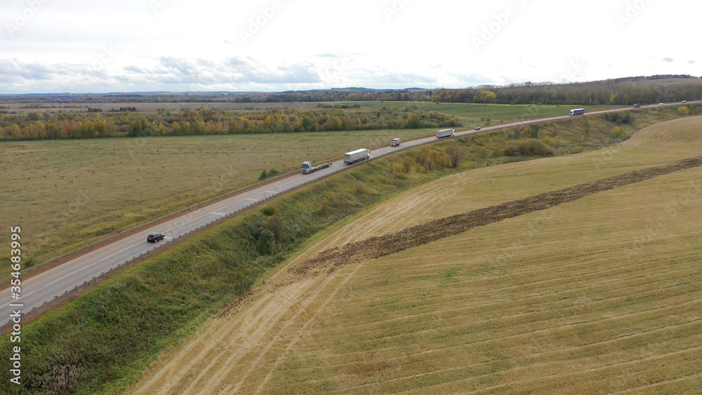 Aerial View of trucks on road driving away in beautiful countryside with green grass in Direction of Loading Warehouse Area. Large Delivery Truck is Moving.