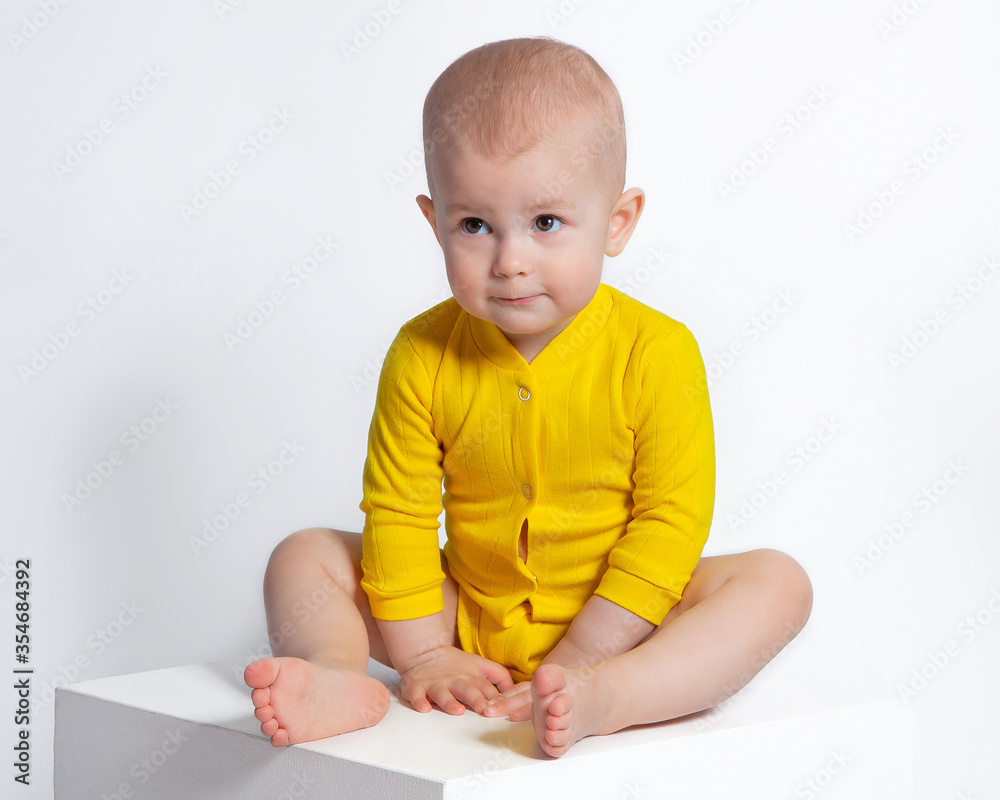 The little girl in the yellow bodysuit looks shy, mysterious, sly. The baby is sitting on a white background. Children's eyes, emotions. The little girl in the yellow bodysuit looks shy, mysterious, sly. The baby is sitting on a white background. Children's eyes, emotions.
