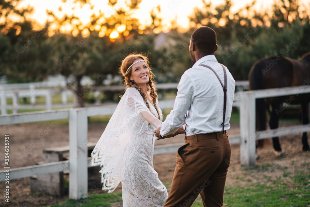 An interracial couple of newlyweds in the style of boho walk on a ranch at sunset. Soft focus.