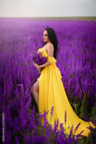 A beautiful young woman in a yellow dress is standing in a blooming purple field. Full-length portrait.