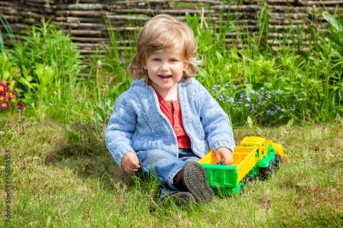 Curly-haired two-year-old blond boy playing in the garden with a tractor. Dacha, nature, vacation, weekend. A family holiday in the fresh air. Flowerbed with flowers, garden decorations.