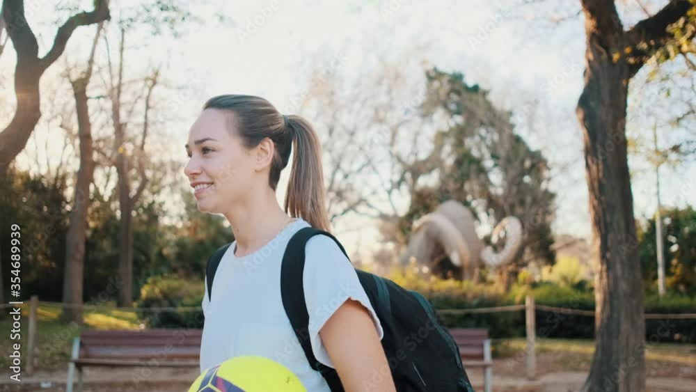 Tracking shot of attractive sporty girl with backpack walking along city park looking in camera playing with a football in hands Tracking shot of attractive sporty girl with backpack walking along city park looking in camera playing with a football in hands