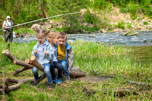 Three children on the Bank of a fast river play, sit on a log and laugh, make faces. Humor, laughter, fun. In the background, a fisherman is fishing.