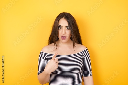 Surprised beautiful female student being in stupor shocked, has astonished expression pointing at herself with finger saying: Who me? Isolated over yellow background.