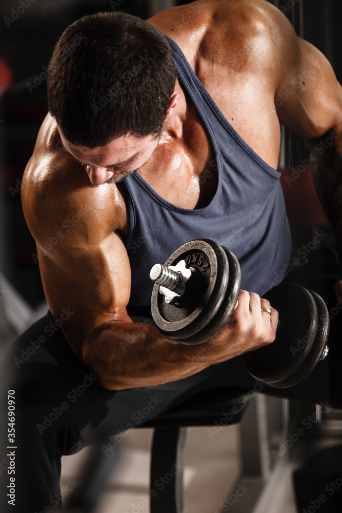 Fitness in gym, sport and healthy lifestyle concept. Handsome athletic man in blue shirt making exercises. Bodybuilder male model training biceps muscles with dumbbell Fitness in gym, sport and healthy lifestyle concept. Handsome athletic man in blue shirt making exercises. Bodybuilder male model training biceps muscles with dumbbell