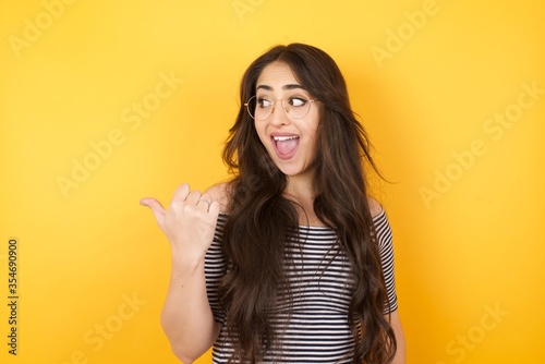 Omg concept. Stupefied caucasian female with surprised expression, opens eyes and mouth widely, points aside with thumb, shows something strange on gray background. Advertisement concept.