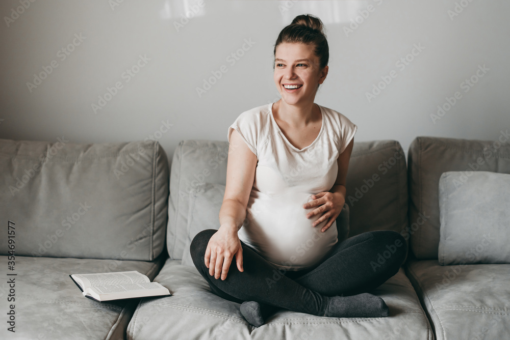 Happy young beautiful pregnant woman sitting on gray couch and reading a book on quarantine at home. Relax, keep calm, rest, coronavirus, virus, isolation, stay at home. Happy young beautiful pregnant woman sitting on gray couch and reading a book on quarantine at home. Relax, keep calm, rest, coronavirus, virus, isolation, stay at home.