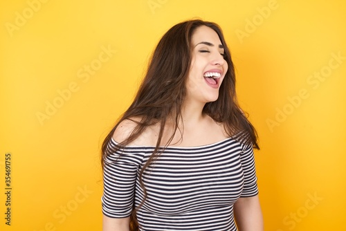 Joyful European woman having fun and laughs at good joke or sings wears casual clothes, standing against gray wall. Happy woman with long hair poses inddoors.