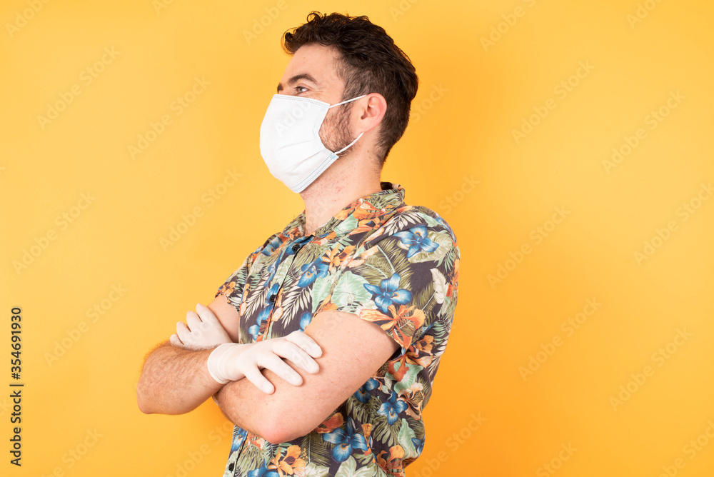 Portrait of cheerful lovely caucasian man crossing arms looking aside having a nice conversation with his friend. Wears hawaiian shirt and medical mask over yellow background Portrait of cheerful lovely caucasian man crossing arms looking aside having a nice conversation with his friend. Wears hawaiian shirt and medical mask over yellow background