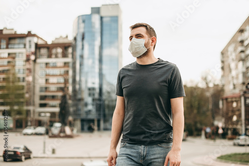 Portrait of brunette european man in a white medical surgical mask, t-shirt in the summer city. Coronavirus, illness, infection, quarantine.