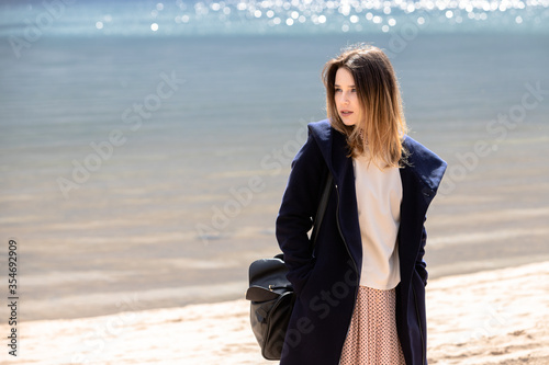 A young adult beautiful girl stands on the shore against the backdrop of an approaching cloud with rain. The woman is wearing a spotty dress  a white sweater and a navy coat.