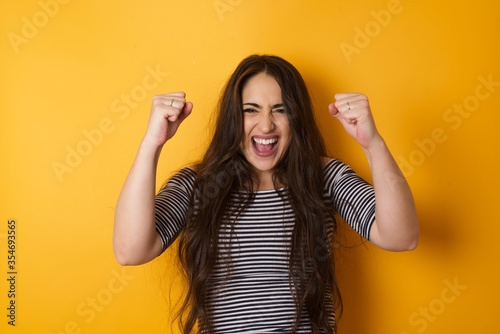 Waist up portrait of strong successful determined young female winner in casual t-shirt raising arms, clenching fists, exclaiming with joy and excitement. Victory, success and achievement concept.