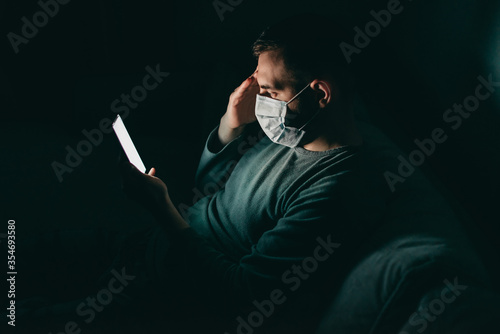 Young white man in medical mask reads news on his phone at home at quarantine at night. Coronavirus, isolation, depression, crisis.