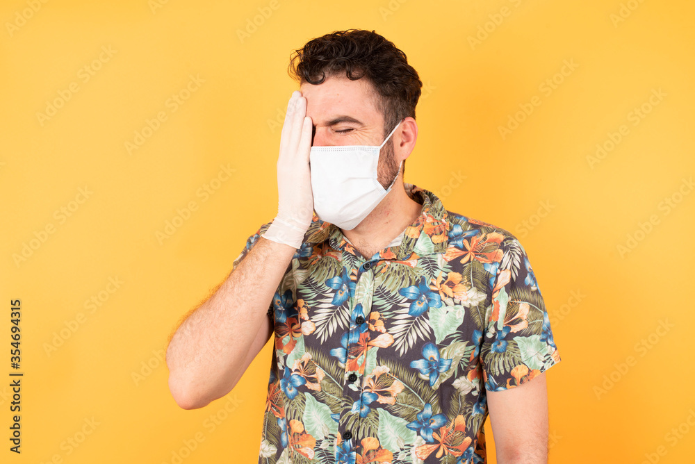 Young caucasian man wearing hawaiian shirt and medical mask standing over yelllow isolated background Yawning tired covering half face, eye and mouth with hand. Face hurts in pain. Young caucasian man wearing hawaiian shirt and medical mask standing over yelllow isolated background Yawning tired covering half face, eye and mouth with hand. Face hurts in pain.
