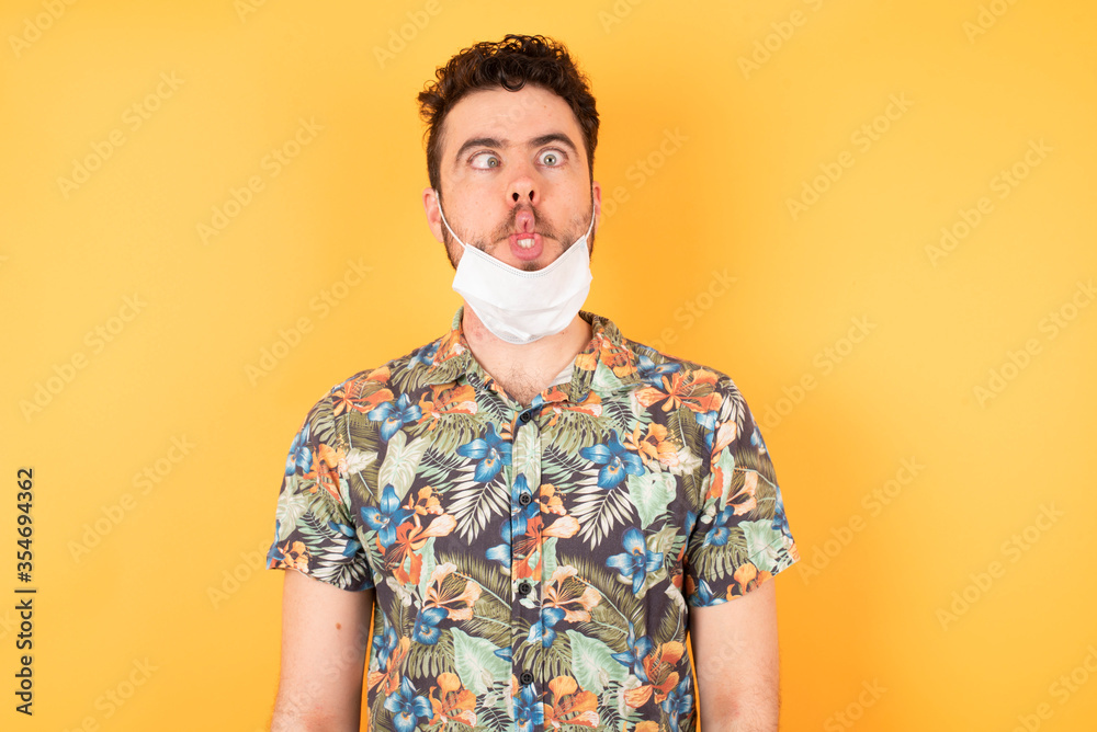 Young caucasian businessman wearing hawaiian shirt and medical mask over isolated yellow background making fish face with lips, crazy and comical gesture. Funny expression. Young caucasian businessman wearing hawaiian shirt and medical mask over isolated yellow background making fish face with lips, crazy and comical gesture. Funny expression.