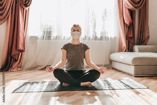 Young beautiful white woman with medical surgical mask doing yoga exercises, meditation. Sport in quarantine at home. Coronavirus, illness, infection, protection, bandage.