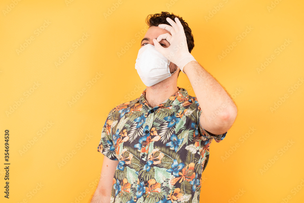 Young caucasian businessman wearing hawaiian shirt and medical mask over isolated yellow background doing ok gesture with hand smiling, eye looking through fingers with happy face. Young caucasian businessman wearing hawaiian shirt and medical mask over isolated yellow background doing ok gesture with hand smiling, eye looking through fingers with happy face.