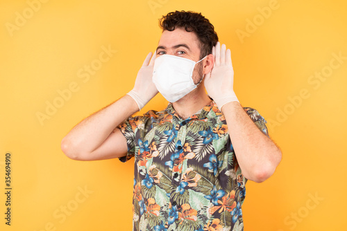 Young caucasian man wearing hawaiian shirt and medical mask  standing over isolated yellow background Trying to hear both hands on ear gesture, curious for gossip. Hearing problem, deaf © Roquillo