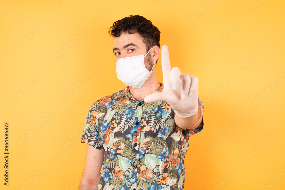 Young handsome businessman wearing hawaiian shirt and medical mask standing over yellow isolated background making fun of people with fingers on forehead doing loser gesture mocking and insulting. Young handsome businessman wearing hawaiian shirt and medical mask standing over yellow isolated background making fun of people with fingers on forehead doing loser gesture mocking and insulting.