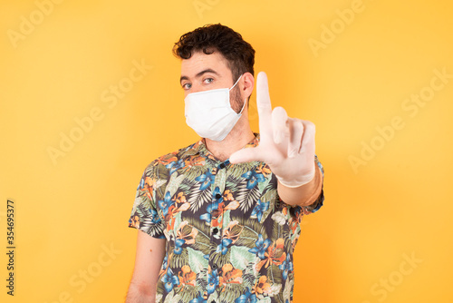 Young handsome businessman wearing hawaiian shirt and medical mask standing over yellow isolated background making fun of people with fingers on forehead doing loser gesture mocking and insulting. © Roquillo