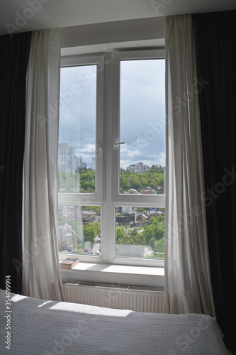 21view of thunderclouds over a park with houses from a white window with black-and-white curtains on a window. white knitted bedspread on a double bed